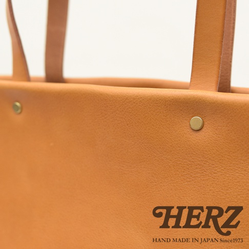 職人手作皮革托特包，呈現溫潤的淺棕色皮革紋理，包身有兩個金屬鉚釘點綴，提把為同色系皮革製作。