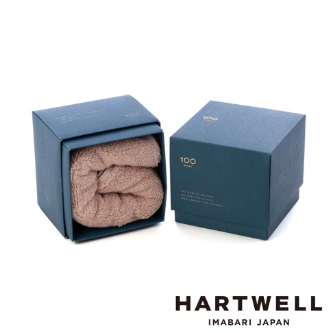 深藍色禮盒裝著一條淺褐色柔軟浴巾，浴巾材質蓬鬆，禮盒上印有金色「100」及日文、英文標語。