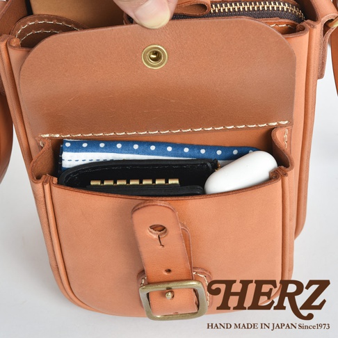 日本職人品牌 HERZ 手作皮革單肩包，以淺棕色硬皮革打造，正面設有復古風格的皮扣翻蓋設計，打開後可見內部收納空間，放置了黑色鑰匙包、白色無線耳機及藍白圓點圖案的布巾，畫面右下方標註 HERZ 品牌字樣與日本手作生產資訊。