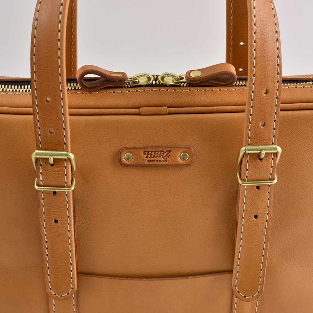 淺棕色皮革製成的手提肩背包，配有金色拉鍊、可調節肩帶和兩個提把。包袋正面中央有一個壓有「HERZ MADE IN JAPAN」字樣的皮革標籤。