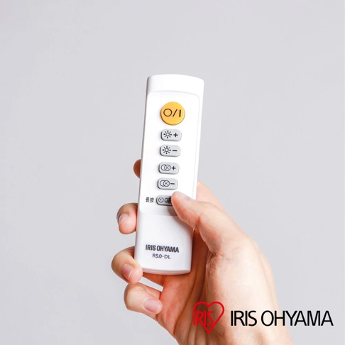 手持白色遙控器，上面有黃色O/I開關按鈕，以及多個用於調整燈光亮度與色溫的按鈕，下方印有IRIS OHYAMA R5.0-DL字樣。