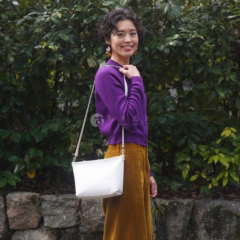 白色皮革手提包，配有細長斜挎肩帶，由一位身穿紫色開襟毛衣和黃色燈芯絨褲的女士展示。