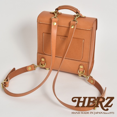 淺棕色皮革學院風迷你後背包，配有金屬扣環和可調節的皮革肩帶，包身正面有兩個方形口袋，頂部有提把，側面有品牌標誌。