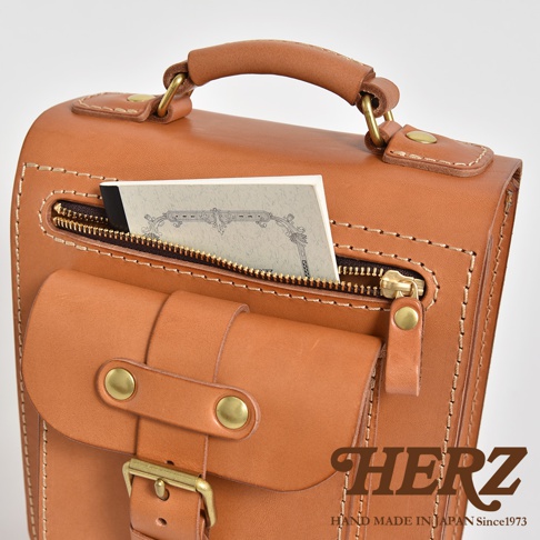 棕色皮革製成的兩用單肩包，有立體前袋和拉鍊開口，袋口露出部分卡片。包包有提把和金屬扣環，車縫線清晰可見。