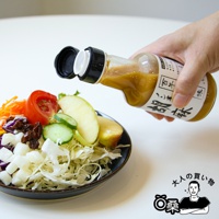 一隻手拿著透明玻璃瓶，瓶內裝有淺棕色的芝麻沙拉醬，瓶身有日文標籤。沙拉醬正從瓶口滴入裝有鮮豔蔬菜和水果的藍色碗中，碗內有高麗菜絲、番茄、小黃瓜片、蘋果片、胡蘿蔔絲和葡萄乾。
