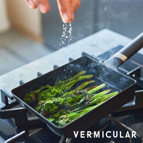 黑色琺瑯鑄鐵玉子燒平底鍋，鍋內有綠色蔬菜正在烹調，鍋蓋未顯示，鍋柄為黑色，鍋身有白色飾環。