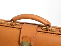 職人手作皮革橫長形單肩包，採用天然皮革材質，呈現溫潤的焦糖棕色。包包頂部設有皮革提把，以黃銅色鉚釘和金屬環固定，展現精緻工藝。