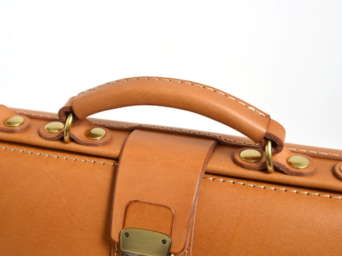 職人手作皮革橫長形單肩包，採用天然皮革材質，呈現溫潤的焦糖棕色。包包頂部設有皮革提把，以黃銅色鉚釘和金屬環固定，展現精緻工藝。