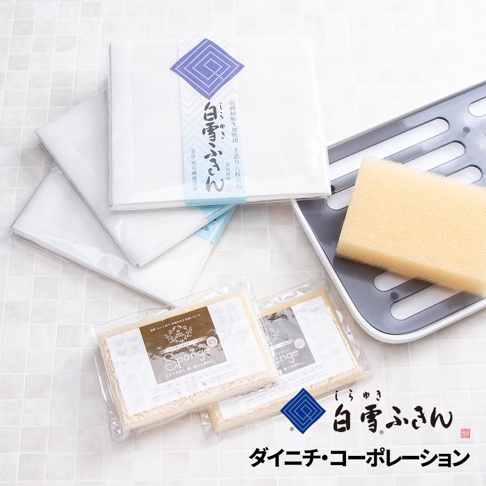 白色網狀清潔布堆疊，其中一張有藍色圖案和日文標籤。旁邊有一個灰色瀝水架，上面放著一塊黃色海綿。下方有兩包透明包裝的白色顆粒狀清潔劑。