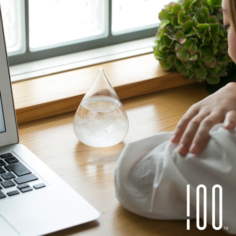 透明玻璃水滴狀天氣瓶，內部裝有白色結晶，置於木質桌面，旁有筆記型電腦與綠色植栽。