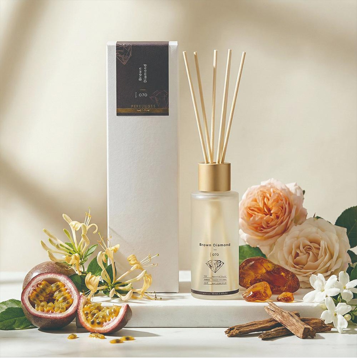 KYARA PERFUMERS 擴香竹，標示著 Brown Diamond 070，搭配著乾燥的香料、百香果、白色花朵和兩朵粉色玫瑰，置於白色大理石檯面上，呈現精緻的居家香氛氛圍。