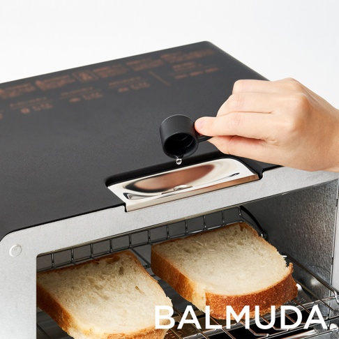 黑色蒸氣烤麵包機，頂部設有注水口，一隻手正用黑色量杯將水倒入。烤麵包機內有兩片烤吐司，放在銀色烤架上。