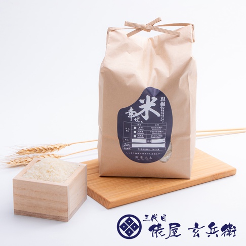 一個印有藍色標籤的米色紙袋，標籤上印有日文文字和圖案，上面有麻繩綁緊的開口。袋子旁邊有一個木製方盒，裡面裝滿了白米。背景是淺色，有幾株麥穗和一個木製砧板。