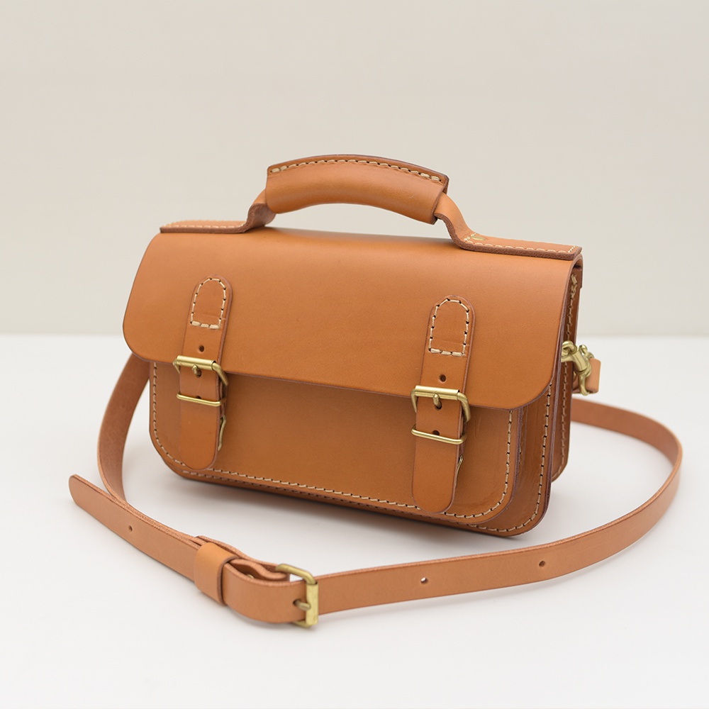 淺棕色皮革迷你肩背包，配有雙金屬扣、頂部提把和可調式斜挎肩帶，邊緣縫線精緻。