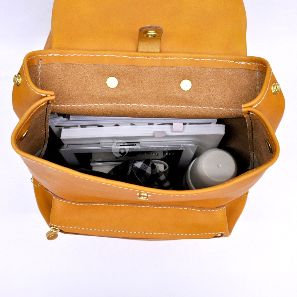 職人手作皮革多口袋後背包 R-135，展示內部空間，裝有書籍、平板電腦、格紋布料及圓筒狀物品。皮革為棕褐色，縫線清晰，配有金屬釦。