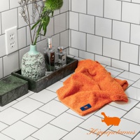 橘色竹棉混紡長毛巾，質地蓬鬆柔軟，帶有深藍色品牌標籤，置於白色磁磚檯面上。