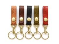 五個職人手作皮革鉤扣鑰匙圈，分別為橘棕色、深棕色、黑色、橄欖綠和紅色，均帶有黃銅色鉤扣和鑰匙環，以及壓印的「HERZ」和「MADE IN JAPAN」字樣。