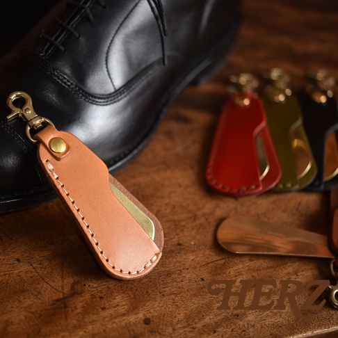 職人手作皮革隨身金屬鞋拔，附有黃銅掛鉤。皮革部分為淺棕色，縫線清晰。背景有紅色、橄欖綠及黑色鞋拔，以及一雙黑色皮鞋。
