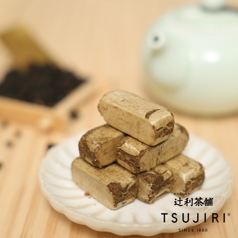 放在白色盤子上的辻利茶舖精選茶酥，呈現堆疊狀，茶酥表面帶有粗糙的紋理與深淺交錯的茶褐色，背景模糊處可見茶具與裝有茶葉的木盒。