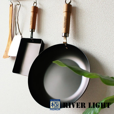 一個黑色不鏽鋼平底鍋，附有木製手柄，掛在牆上，旁邊有一個方形平底鍋和一個木製鍋鏟。