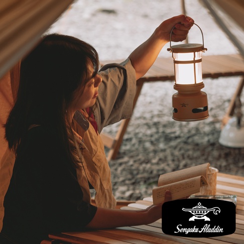 一位女性在帳篷內手持一個黃色神燈造型的音箱，音箱散發溫暖光芒，她正閱讀一本打開的書，桌上還有其他物品。