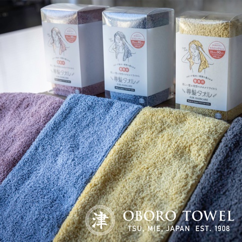 展現四款不同顏色的髮擦巾，包含粉紫色、淺藍色、鵝黃色和深灰色，材質為柔軟絨毛，產品包裝盒上印有品牌標誌和產品說明。