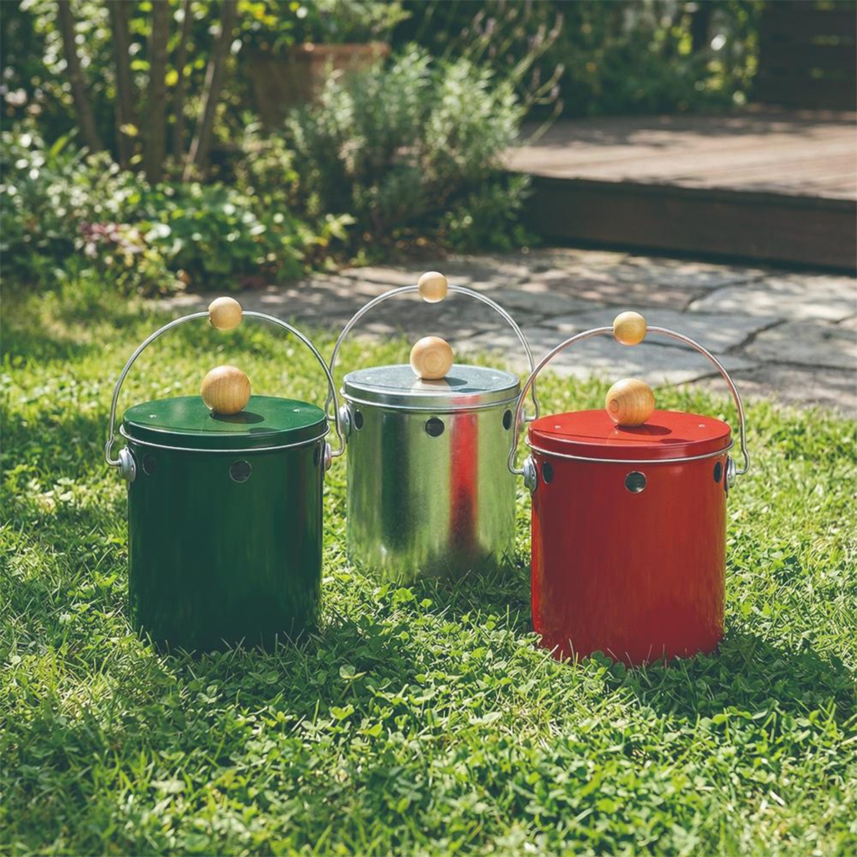 三款OBAKETSU手工製蚊香桶，分別為綠色、銀色和紅色，放置於草地上，背景是模糊的綠色植物和木質地板。