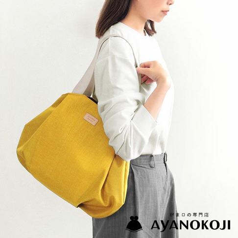 一位女士身著白色上衣和灰色條紋長褲，肩背一個亮黃色的托特包，包上有名為 AYANOKOJI 的日文品牌標誌。