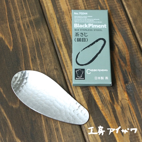 一個銀色不鏽鋼製成、帶有錘紋的茶勺，置於木紋背景上，旁邊是產品包裝盒。