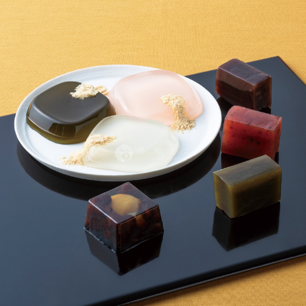 白色盤子上放著三個半透明的果凍，表面灑有細緻的黃豆粉，盤子旁放置著數個切成長方體與正方體的羊羹，包含紅豆、栗子、抹茶等多種色澤與口味，整體擺放在黑色托盤上，背景為簡約的黃色布紋。