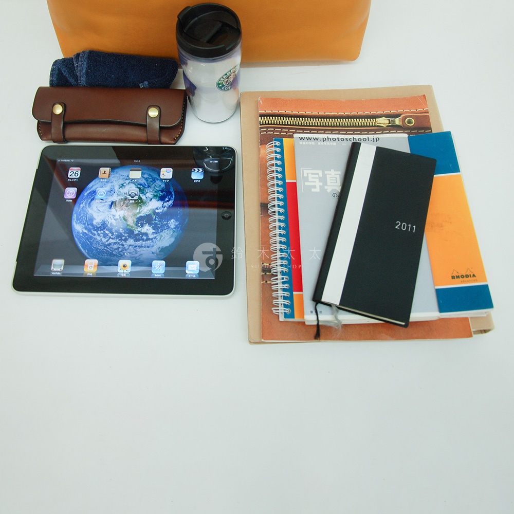 皮革軟式杜勒斯包，棕色，附有金色金屬扣，旁邊是深藍色牛仔布、白色馬克杯、平板電腦和幾本筆記本。