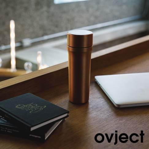 銅色金屬材質的三用保溫瓶，瓶身光滑，頂部有銀色蓋子，旁邊放著黑色筆記本和白色筆記型電腦，背景是模糊的窗戶和燈光。