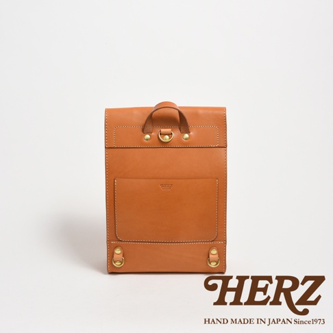 棕色皮革製作的紙袋風後背包，正面有貼袋和品牌標誌，頂部有提把和金屬扣環，底部有兩個金屬環。