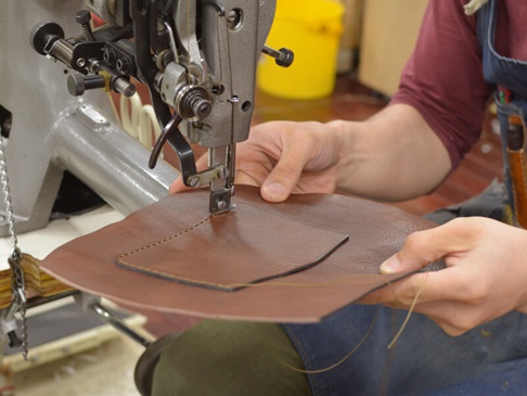 職人手作皮革屋瓦風迷你後背包 (R-6) 的製程，特寫皮革匠人使用工業縫紉機縫製棕色皮革的細節，皮革表面光滑，有精緻的縫線與貼袋，展現高品質手工藝。