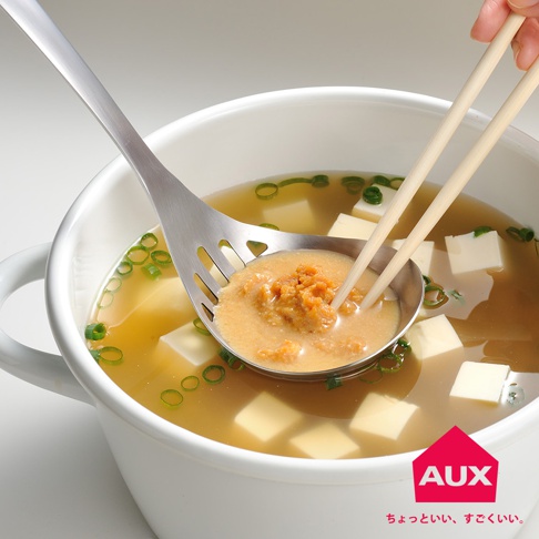銀色金屬濾湯火鍋杓，杓頭有長方形濾孔，正盛著味噌湯與豆腐塊，湯內有蔥花。一雙淺木色筷子夾著味噌塊，伸入杓頭。