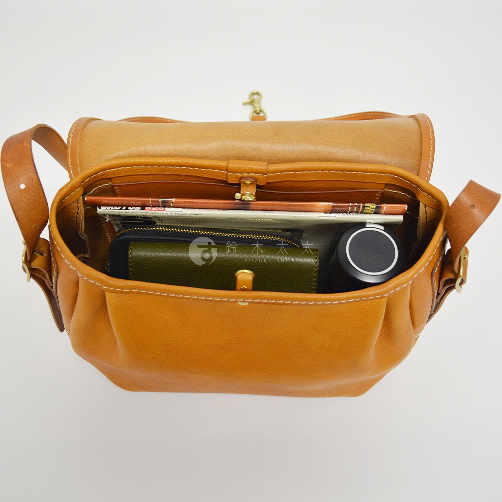職人手作皮革復古翻蓋郵差包，打開的包內裝有雜誌、綠色皮夾和黑色水杯，包身為棕褐色皮革材質，車縫線明顯。