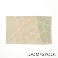 一條淺米色與淺綠色拼接的純棉浴巾，表面印有白色幾何線條圖案，右下角有一個品牌標籤。