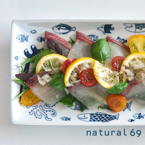 白色長方餐盤，盤緣飾有藍色海洋生物圖案，盤中盛有生魚片、檸檬片、小番茄及綠葉裝飾，整體呈現日式料理風格。