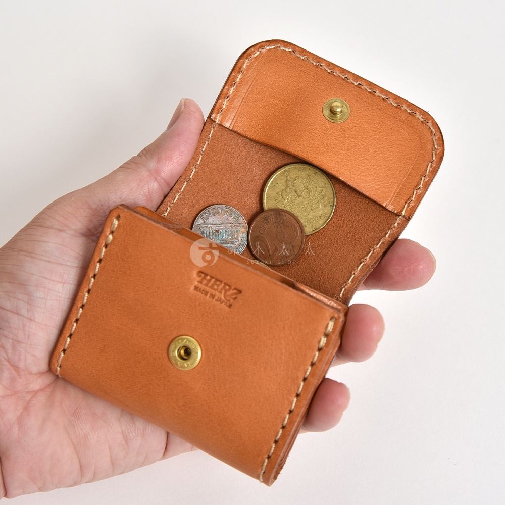 一隻手持著日本HERZ品牌的淺棕色真皮零錢包，零錢包呈現雙折結構，表面有細緻的手工縫線，中間壓印著品牌標誌，內部裝有幾枚金屬硬幣，整體風格簡約且充滿職人手作質感。