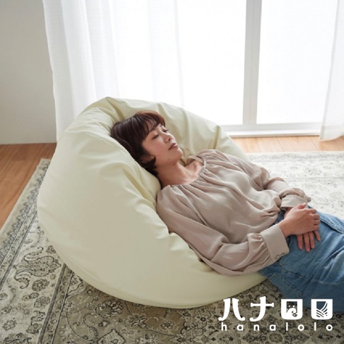 淺黃色皮革材質的懶骨頭沙發，造型如同洋蔥，一位女性躺臥其中，沙發置於印花地毯上，背景為窗戶與窗簾。