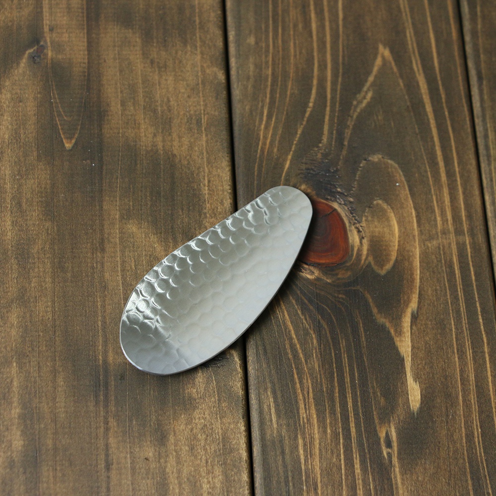 一個橢圓形的不鏽鋼茶勺，表面有錘紋紋理，置於深棕色木紋桌面上。