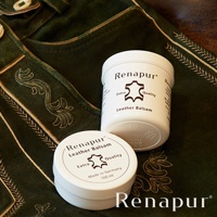 兩罐白色 Renapur 皮革保養油，標籤上印有品牌名稱、皮革圖示和「Leather Balsam」字樣，置於深綠色絨面皮革製品上，製品飾有精緻的金色刺繡。