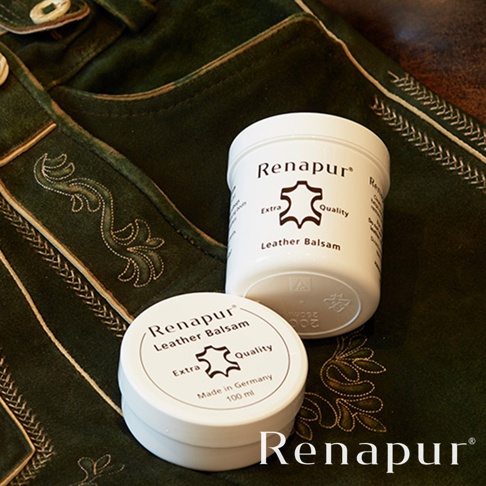 兩罐白色 Renapur 皮革保養油，標籤上印有品牌名稱、皮革圖示和「Leather Balsam」字樣，置於深綠色絨面皮革製品上，製品飾有精緻的金色刺繡。