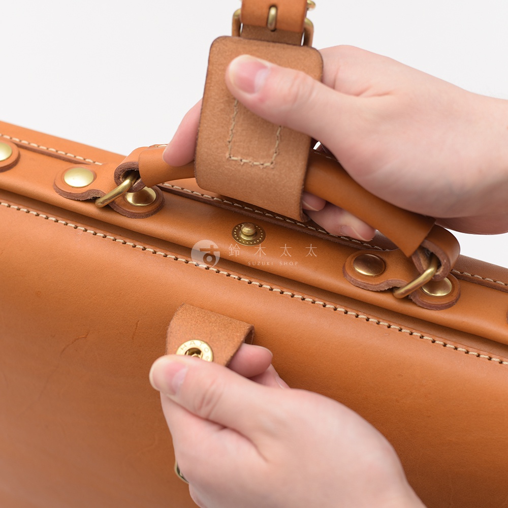 職人手作皮革軟式杜勒斯包，展示棕褐色皮革材質，有精緻縫線與金屬釦件，雙手正在操作包款提把與釦帶。
