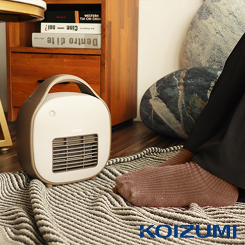 米白色及淺棕色圓角方形陶瓷電暖器，頂部有提把，正面有出風口及指示燈，放置於條紋地毯上，旁有木製櫃子與裝飾品，以及穿著棕色襪子的人的腳。