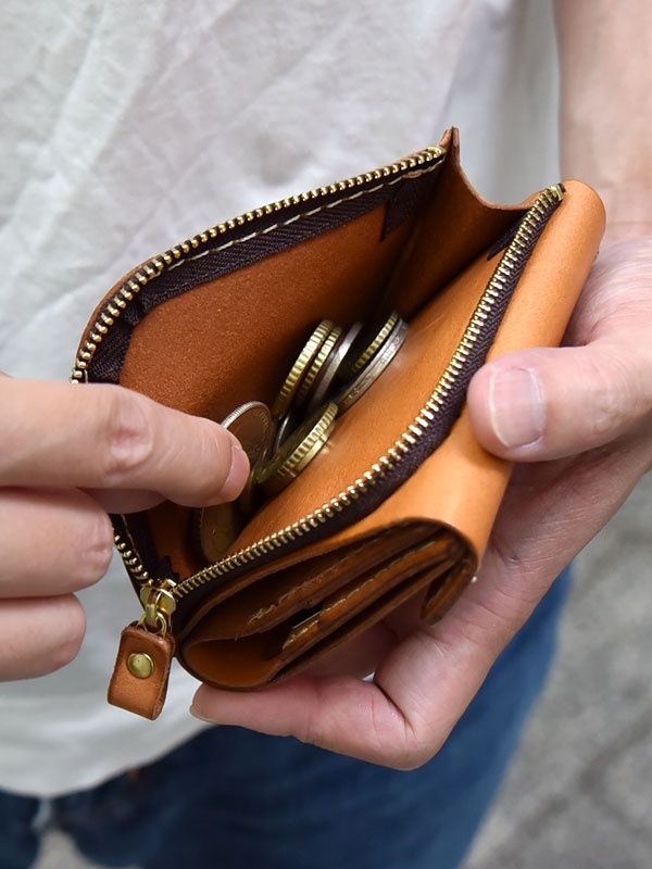 一隻手正在打開一個棕色皮革方形短夾，露出內部裝有硬幣的拉鍊口袋。拉鍊為黃銅色，帶有一個皮革拉環。
