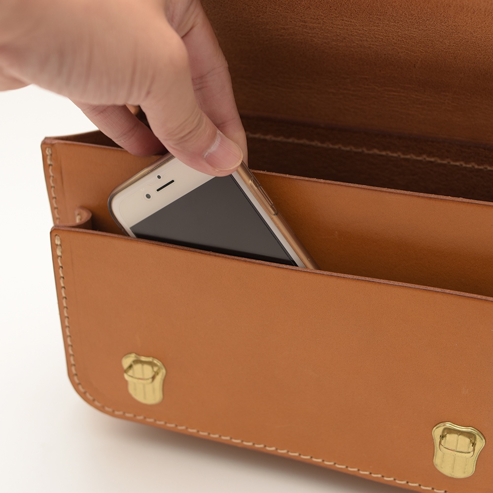 職人手作皮革迷你肩背包，展示側面，將手機放入包款內袋。背包為淺棕色皮革材質，有雙扣設計，縫線清晰。