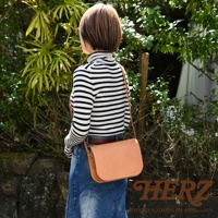 一位女性背著一個淺棕色皮革翻蓋橫式單肩包，包包有明顯縫線細節，搭配條紋高領衫和牛仔裙。