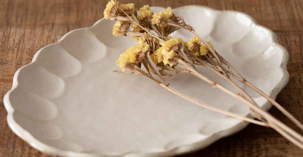 六魯粉引花型橢圓料理盤，米白色陶瓷材質，邊緣有花瓣狀凹凸紋理，盤面有乾燥黃花束裝飾。