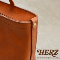 HERZ 品牌標誌，下方有 HAND MADE IN JAPAN Since1973 字樣，圖片右側為手提包的皮革邊緣細節，以特寫方式呈現。